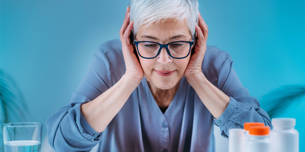Person mit grauem Haar sitzt an einem Tisch und hält beide Hände an den Kopf; im Vordergrund stehen ein Glas Wasser und mehrere Medikamentenbehälter.